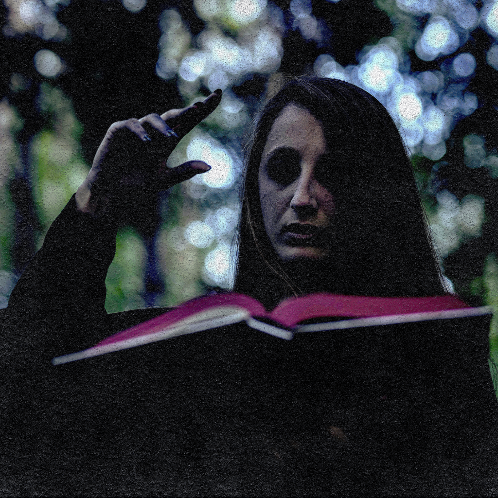 A young woman sits alone in a dark forest, reading from a book with a dark gothic cover. The book is full of dark fiction, with detailed and gruesome illustrations. It is easy to imagine her sitting there, engrossed in this dark fantasy world, surrounded by the real-world dark forests at night.